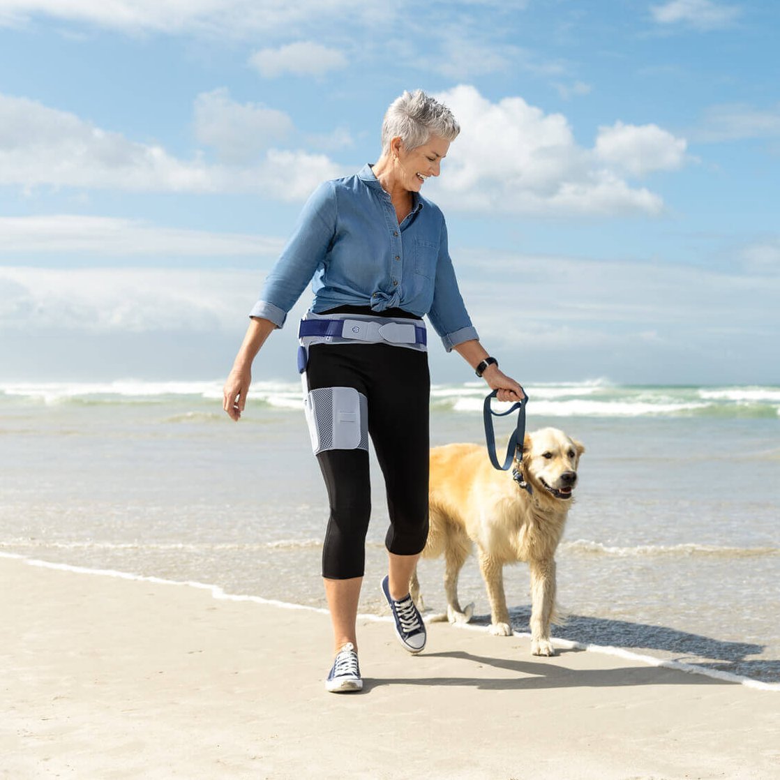 Das Bild zeigt eine ältere Frau beim Strandspaziergang mit ihrem Hund. Die Frau trägt eine CoxaTrain Orthese, die speziell für Hüftarthrose-Patienten entwickelt wurde.