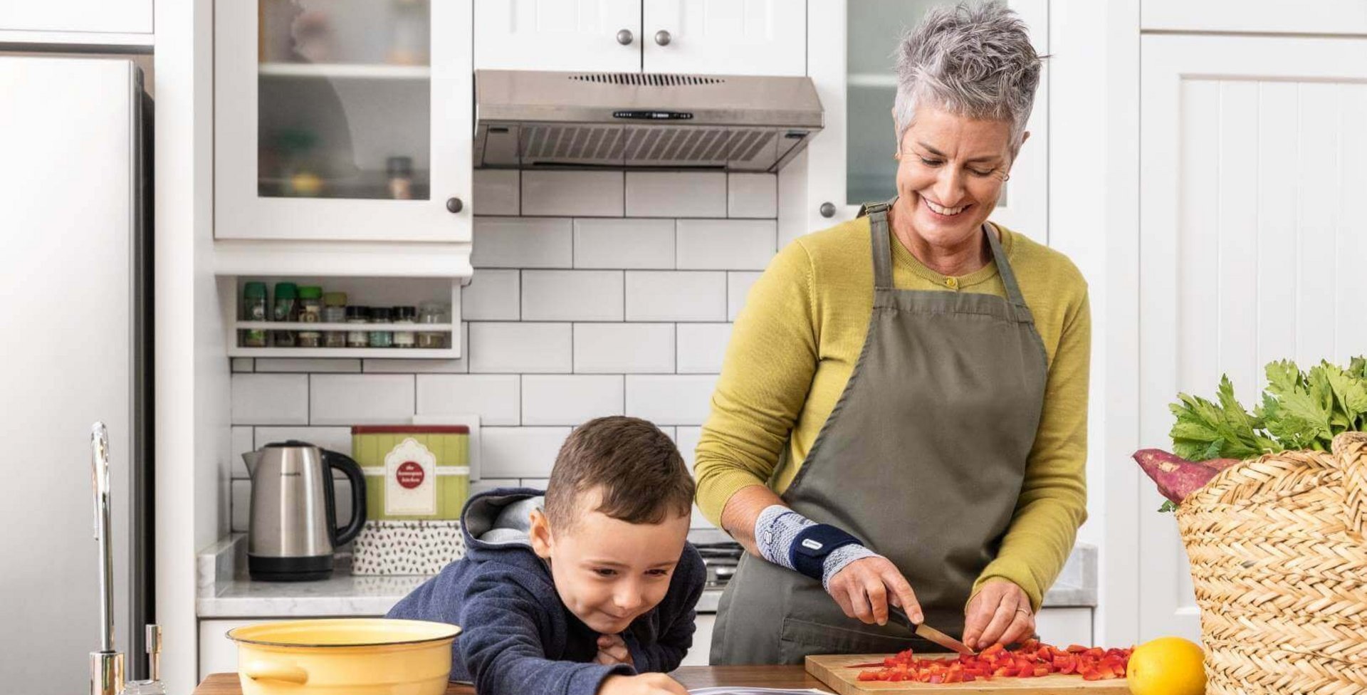 Das Bild zeigt eine ältere Frau in einer Küche, die gemeinsam mit ihrem Enkel kocht. Die Dame trägt eine ManuTrain Bandage zur konservativen Therapie ihrer Handgelenksarthrose.