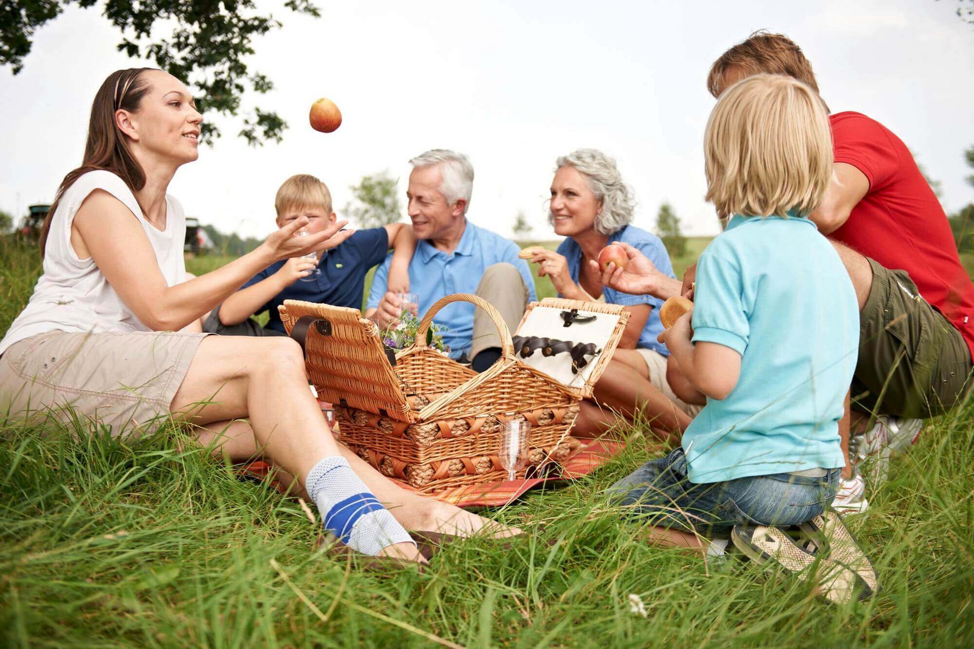 Eine Großfamilie von sechs Personen sitzt auf einer Wiese. In der Mitte befindet sich ein Picknickkorb. Die Frau an der linken Seite wirft einen Apfel in die Höhe. Sie trägt eine stabilisierende Sprunggelenksbandage von Bauerfeind am rechten Knie.  
