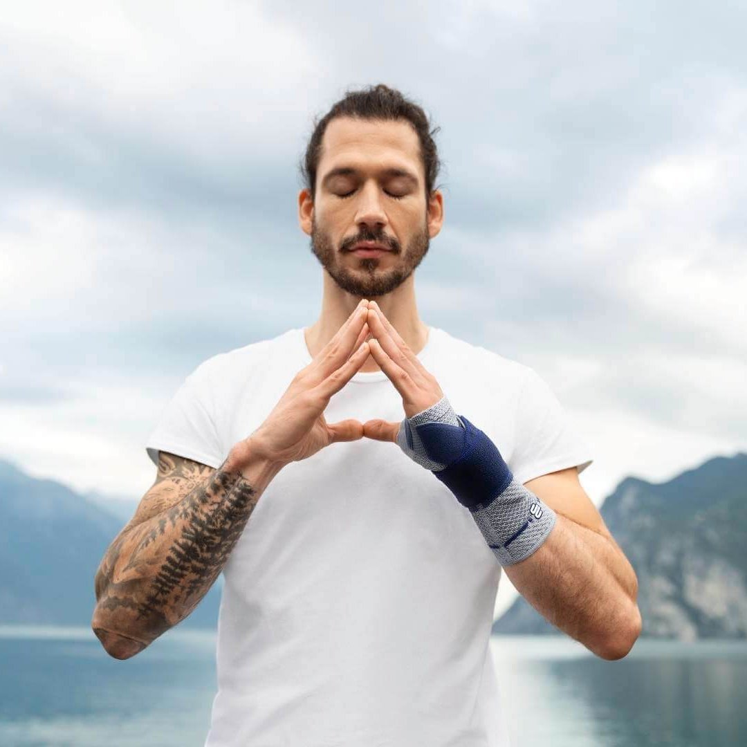Auf dem Bild ist ein junger Mann an einem See zu sehen, der eine Yoga-Pose einnimmt und dabei eine ManuTrain Handgelenkbandage trägt, um Schmerzen im Handgelenk zu lindern.