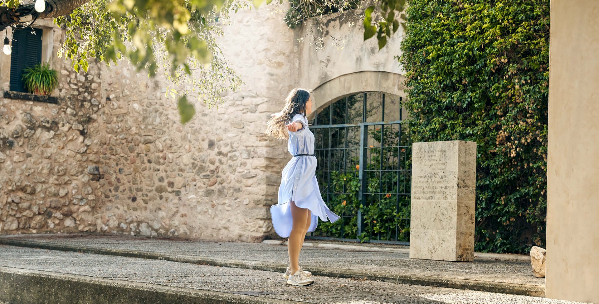 Eine junge Frau in einem fließenden hellblauen Kleid dreht sich mit ausgebreiteten Armen auf einer Steinterrasse vor einer mediterranen Steinmauer. Sie trägt dezente VenoTrain Kompressionsstrümpfe, die sich harmonisch in das Outfit einfügen.