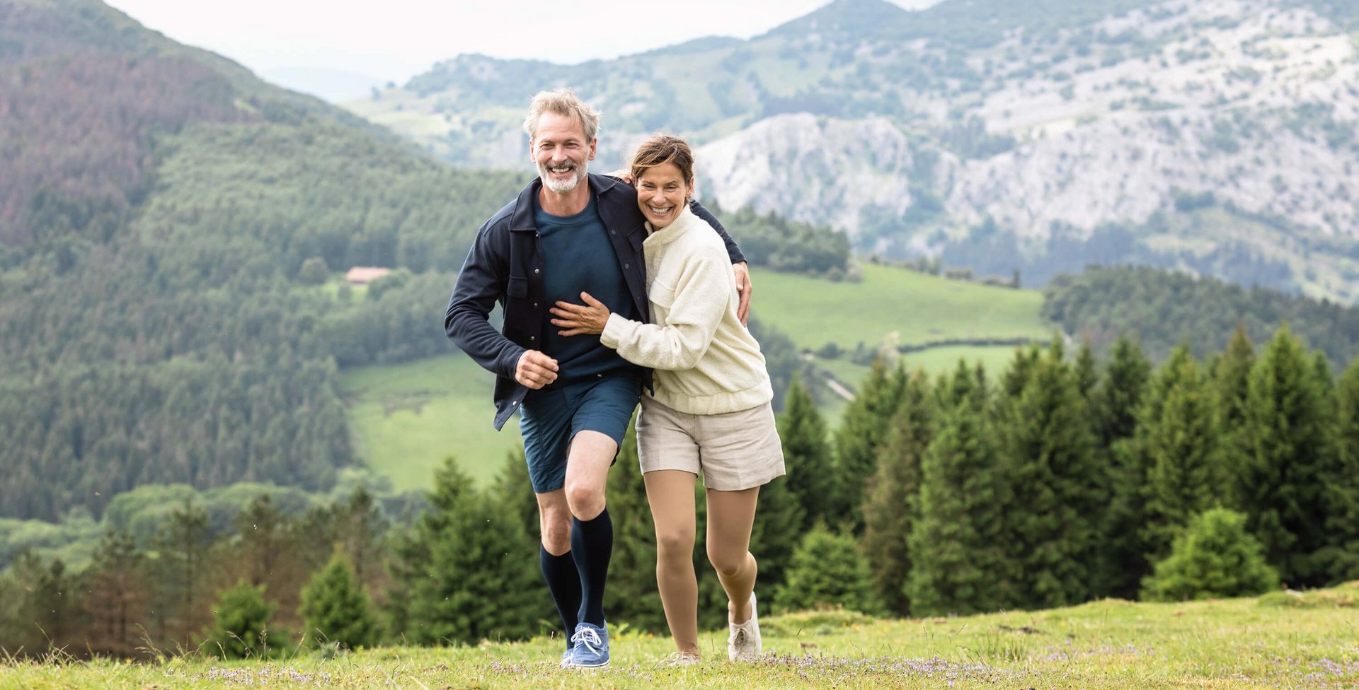 Ein Mann und eine Frau laufen Arm in Arm in einer Berglandschaft über eine Wiese. Beide tragen VenoTrain impuls Kompressionsstrümpfe von Bauerfeind. Diese helfen bei Besenreisern.