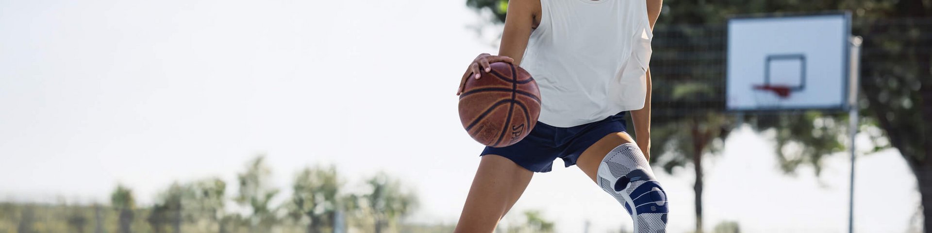 Junge Frau mit weissem Trägershirt, dunkelblauer Sporthose und weisser Kniebandage spielt Basketball auf einem Aussenplatz mit blauem Bodenbelag; sie dribbelt den Ball und lächelt in die Kamera, im Hintergrund ist ein Basketballkorb und Bäume sichtbar.
