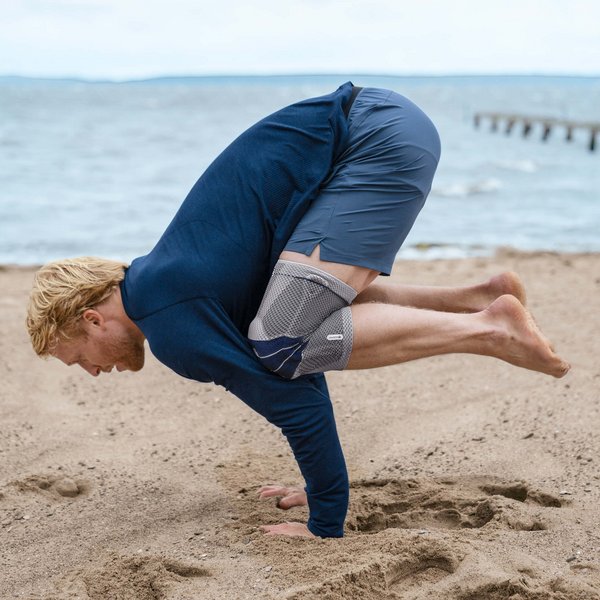 Das Bild zeigt einen Mann in Yoga-Pose am Strand. Am rechten Knie trägt er eine GenuTrain zur Unterstützung. Die Bandage bietet u.a. Unterstützung bei einer Kniegelenkinstabilität.