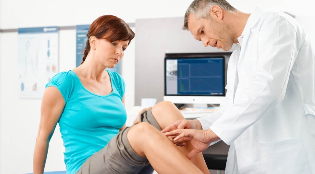 Das Bild zeigt eine junge Frau auf einer Liege in einem Behandlungszimmer. Der Arzt untersucht ihre Knie, um die Ursachen sowie eine optimale Behandlung der Knieschmerzen zu identifizieren.