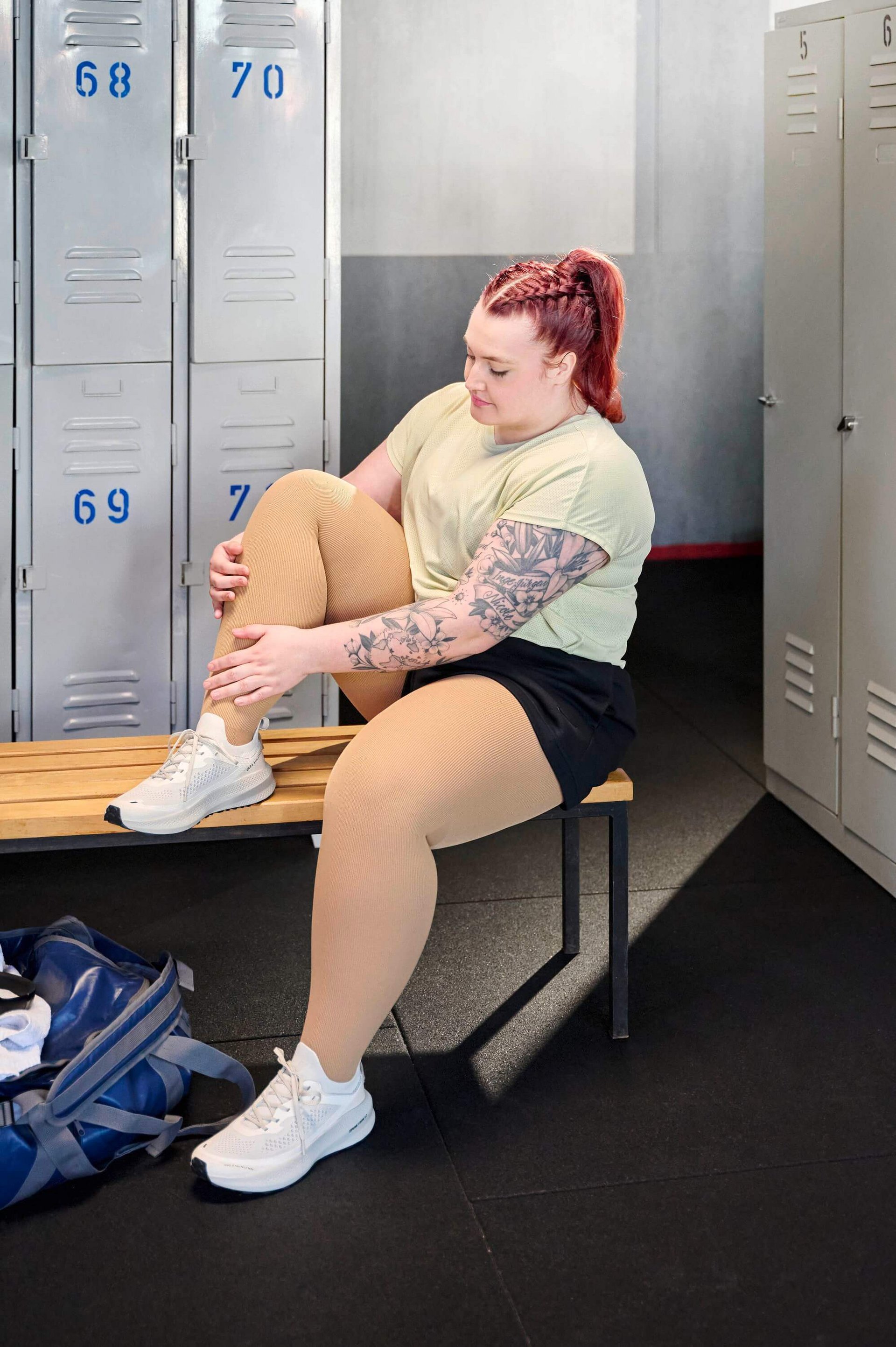 Person sitzt auf einer Holzbank in einer Umkleidekabine und hält ihr angewinkeltes Bein, das mit cremefarbenen Kompressionsstrümpfen bekleidet ist. Sie trägt ein helles Sportshirt, schwarze Shorts und weisse Sportschuhe. Im Hintergrund graue Spinde mit blauen Nummern.