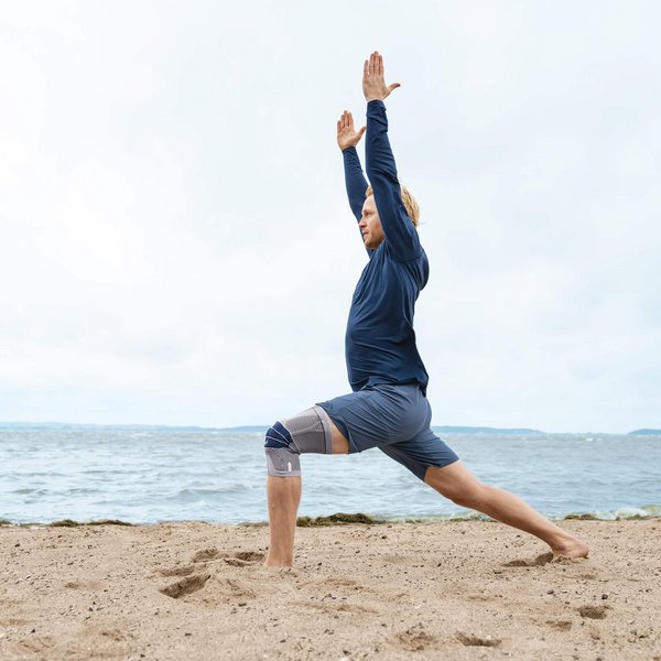 Das Bild zeigt einen Mann in Yoga-Pose am Strand. Am rechten Knie trägt er eine GenuTrain zur Unterstützung. Die Bandage bietet u.a. Unterstützung bei einem Kniegelenkserguss.
