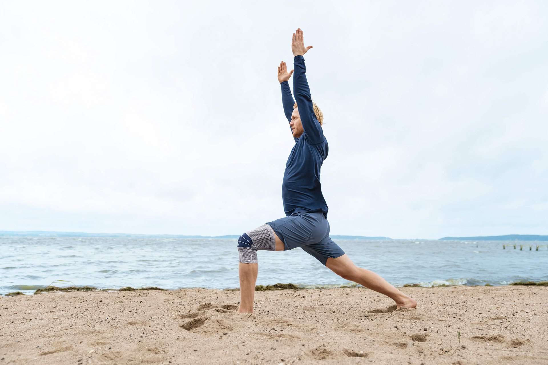 Das Bild zeigt einen Mann in Yoga-Pose am Strand. Am rechten Knie trägt er eine GenuTrain zur Unterstützung. Die Bandage bietet u.a. Unterstützung bei einem Kniegelenkserguss.