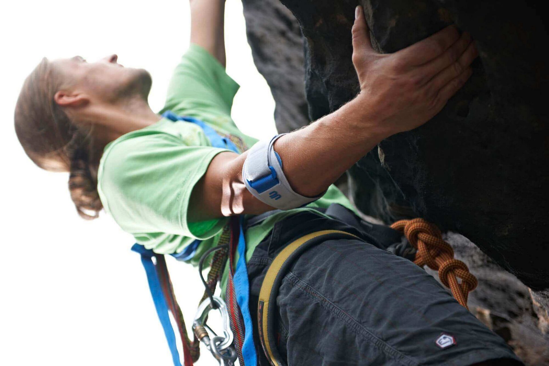  Ein junger Mann mit Sicherung klettert an einem Felsen. Er trägt eine EpiPoint von Bauerfeind am rechten Arm, eine schmerzlindernde Epicondylitis-Spange für den Golfarm.
