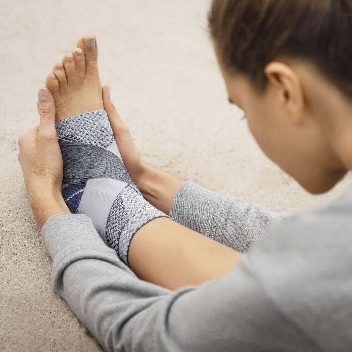 Das Bild zeigt eine Frau, die am Strand in einer Yoga-Pose steht. Sie trägt eine MalleoTrain S Plus Sprunggelenkbandage von Bauerfeind, um ihr Sprunggelenk zu unterstützen.
