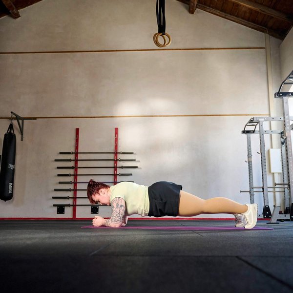 Person macht eine Unterarmstütz-Übung (Plank) auf einer Fitnessmatte in einem geräumigen Trainingsraum mit hohen Decken, Hantelstangen an der Wand und Fenstern auf der rechten Seite.