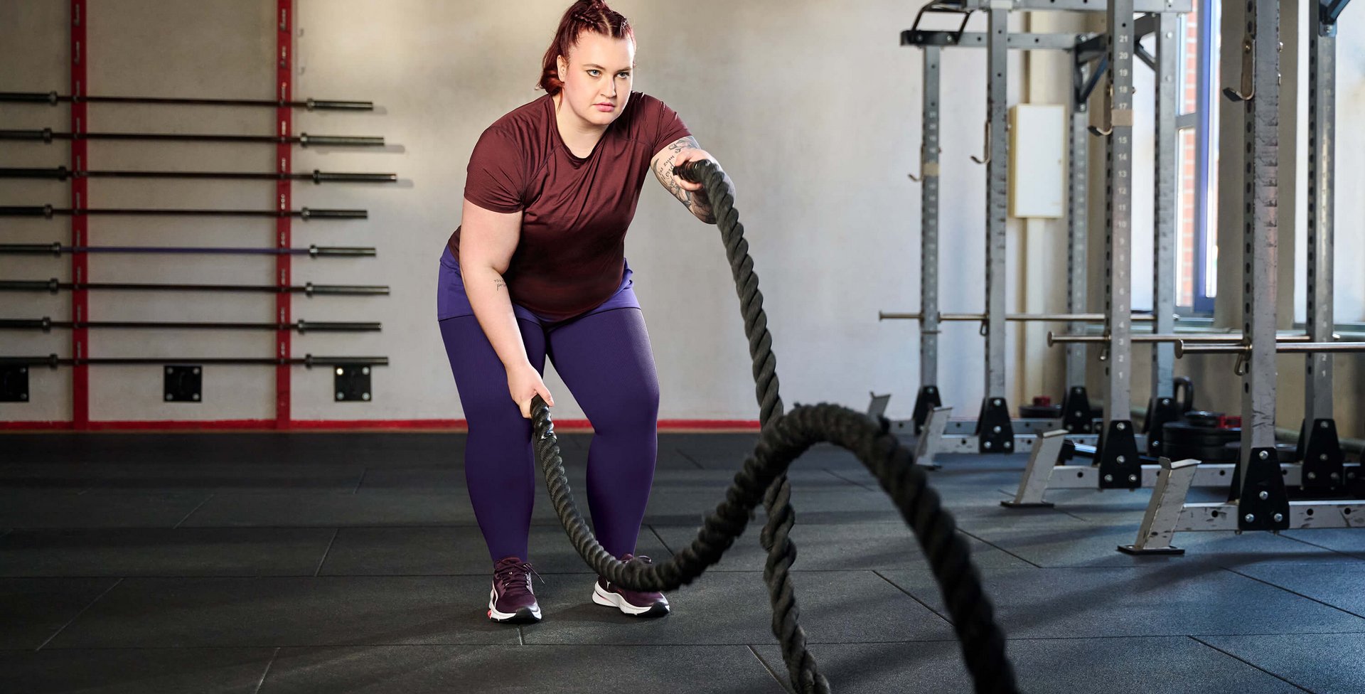 Eine Frau trainiert in einem Fitnessstudio mit Battle Ropes. Sie trägt ein bordeauxfarbenes T-Shirt, lila Leggings und Sportschuhe. Im Hintergrund sind Hanteln an der Wand sowie Krafttrainingsgeräte zu sehen.