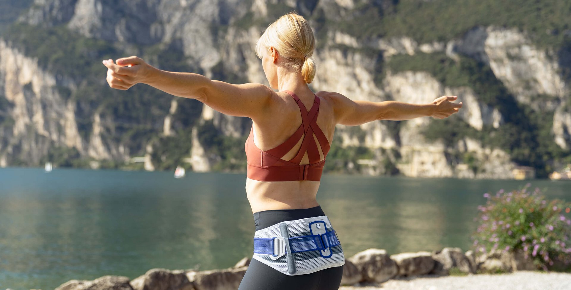 Rückansicht einer Frau mit der SacroLoc Rückenorthese, die ihre Arme ausbreitet. Sie trägt ein rotes Sporttop und dunkle Leggings und steht vor einer malerischen See- und Berglandschaft.