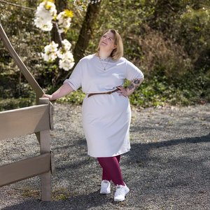 Frau steht draussen auf einem Kiesweg neben einem Holzzaun und blickt nach oben zu blühenden weissen Kirschblüten. Sie trägt ein weisses knielanges Kleid mit braunem Gürtel, bordeauxrote Leggings und weisse Turnschuhe. Im Hintergrund sind Bäume und Sträucher zu sehen, die Frühlingsstimmung vermitteln.