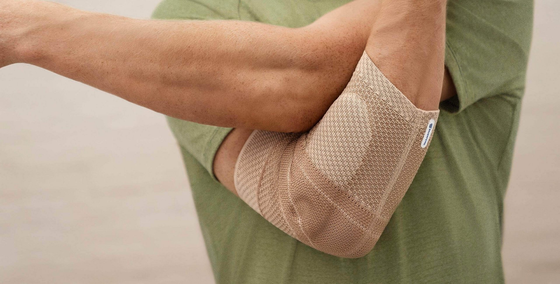 Das Bild zeigt einen Ausschnitt einer Person mit einem grünen T-Shirt, die Armdehnungsübungen gegen einen Tennisarm durchführt. Die Person trägt eine schmerzlindernde Ellenbogenbandage namens EpiTrain von Bauerfeind, die in der Farbe Beige erhältlich ist.