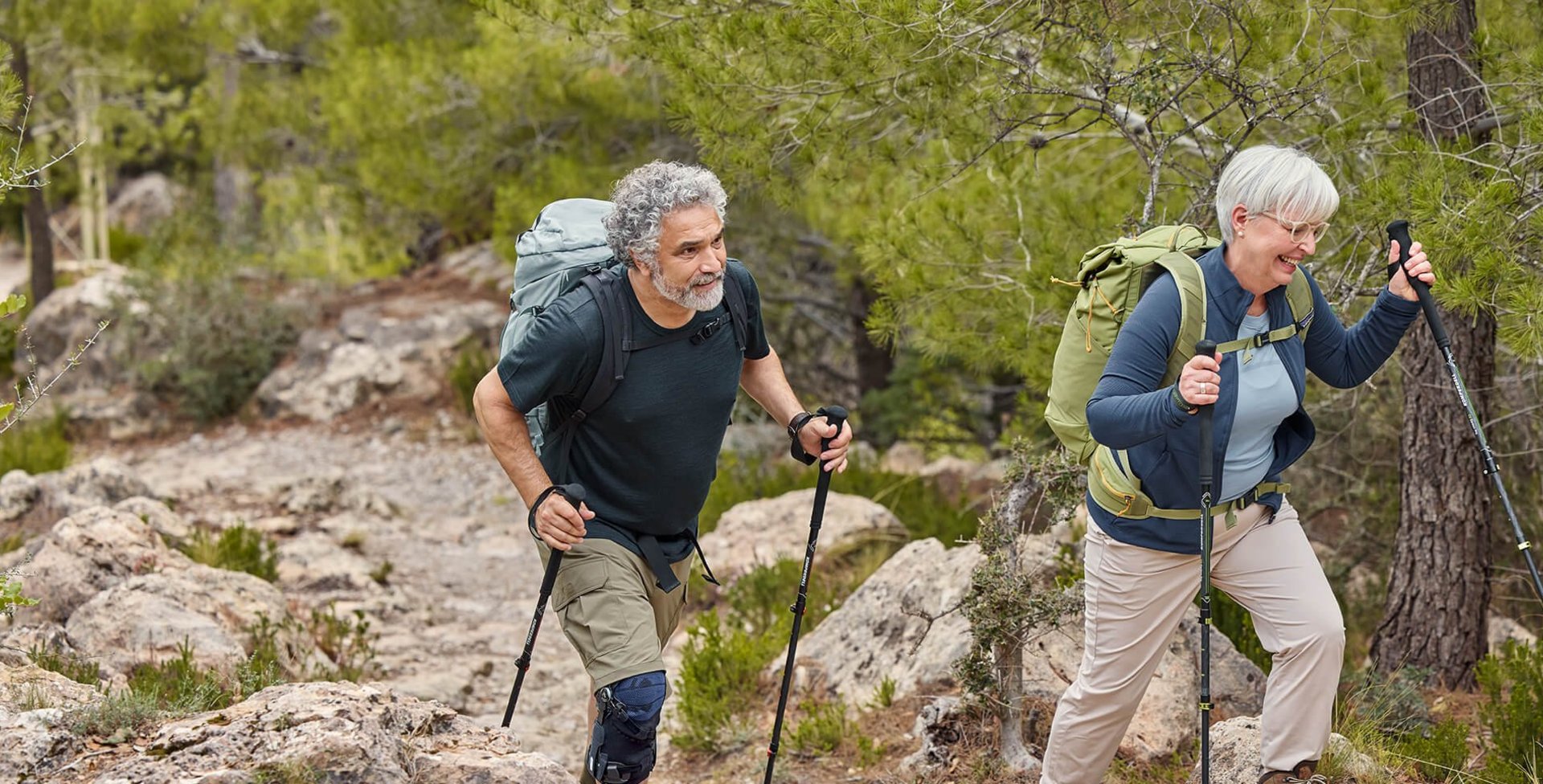 Ein Mann und eine Frau wandern auf einem steinigen Weg durch einen Wald. Beide tragen Wanderausrüstung mit Rucksäcken und Wanderstöcken. Der Mann trägt eine GenuTrain OA Knieorthese am rechten Knie, die seine Bewegung beim Anstieg unterstützt.