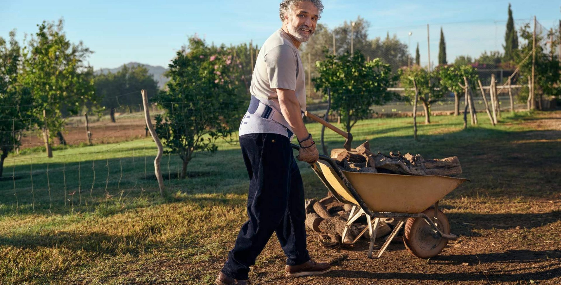 Ein älterer Mann mit grauen Locken schiebt eine Schubkarre voller Holz auf einem ländlichen Grundstück mit Bäumen und einem Zaun. Er trägt eine Lumboloc Forte Rückenorthese zur Unterstützung des unteren Rückens sowie bequeme Kleidung und feste Schuhe. 