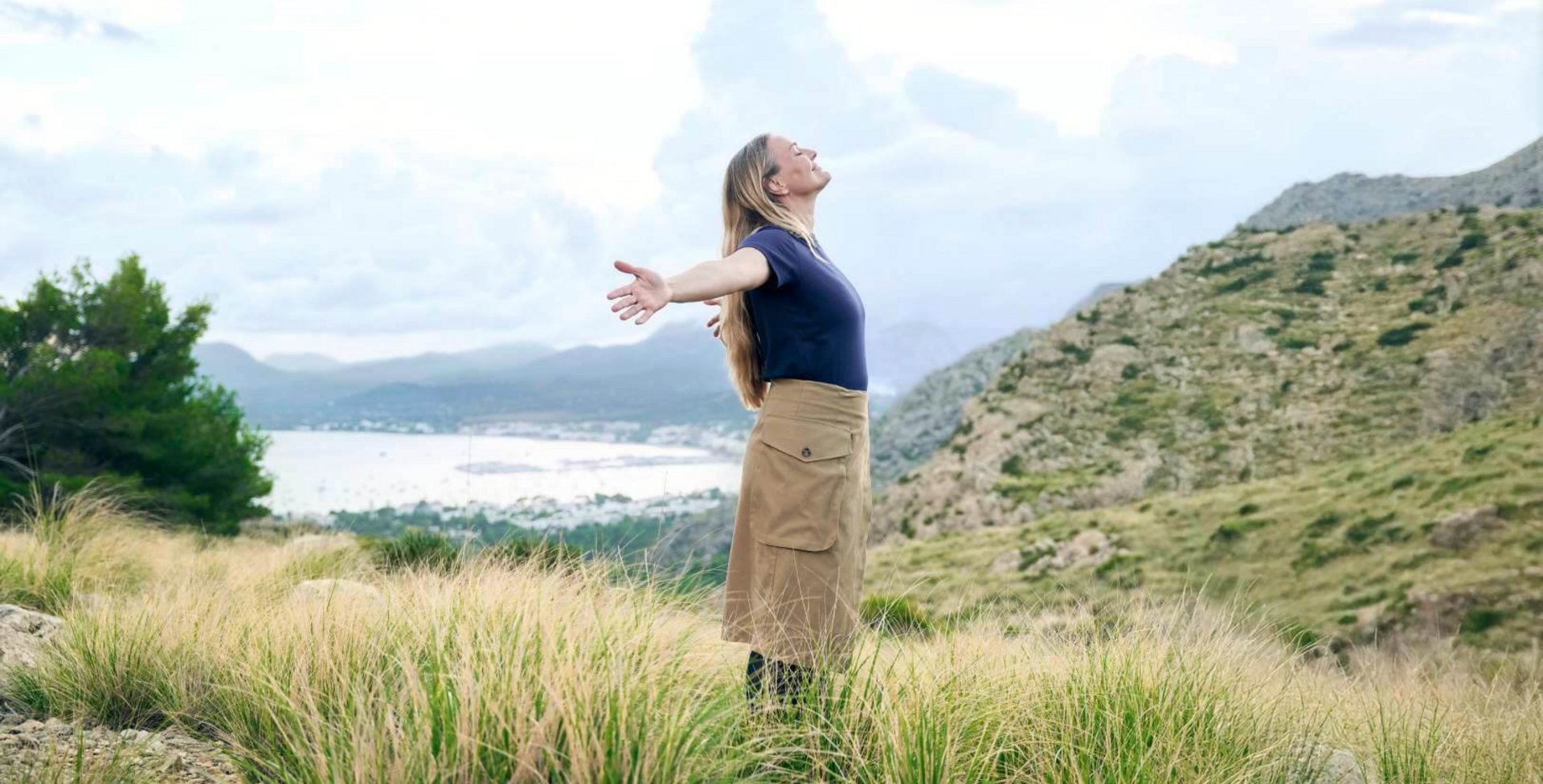 Eine Frau mit langen, offenen Haaren steht auf einem Hügel mit Blick auf eine malerische Küstenlandschaft. Sie breitet die Arme aus, atmet tief ein und genießt die frische Bergluft. Ihr Outfit in Erdtönen ergänzt den natürlichen Look, während dezente VenoTrain Kompressionsstrümpfe für Komfort sorgen.