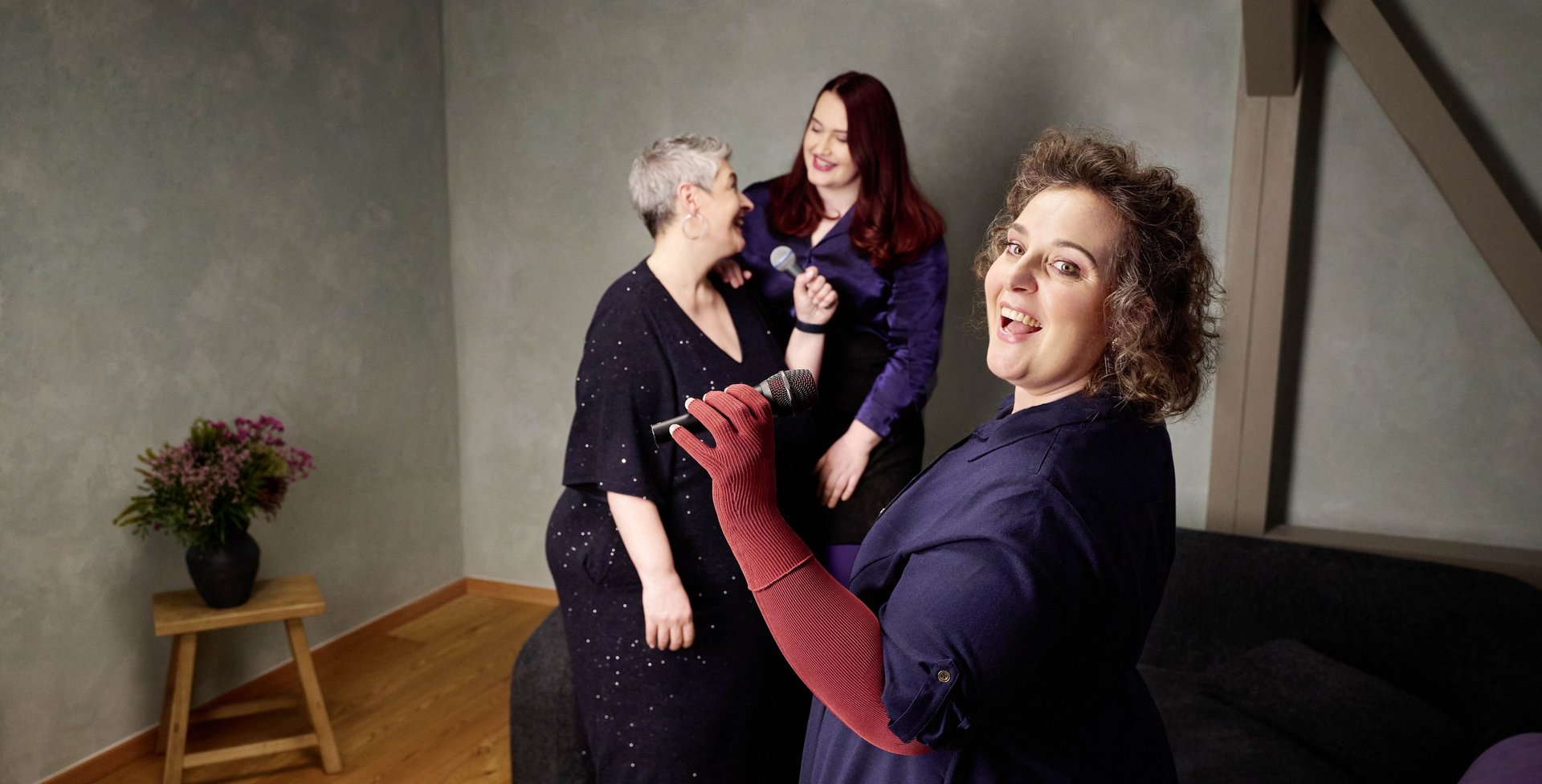 Drei Frauen stehen in einem Wohnzimmer mit hellgrüner Wand und singen gemeinsam mit Mikrofonen. Eine Frau im Vordergrund trägt dunkelblaues Kleid und rote Kompressionshandschuhe, während sie lächelnd in die Kamera singt. Im Hintergrund lachen zwei weitere Frauen, eine mit kurzen grauen Haaren und eine mit langen roten Haaren. Links im Bild steht ein Hocker mit einer Blumenvase.
