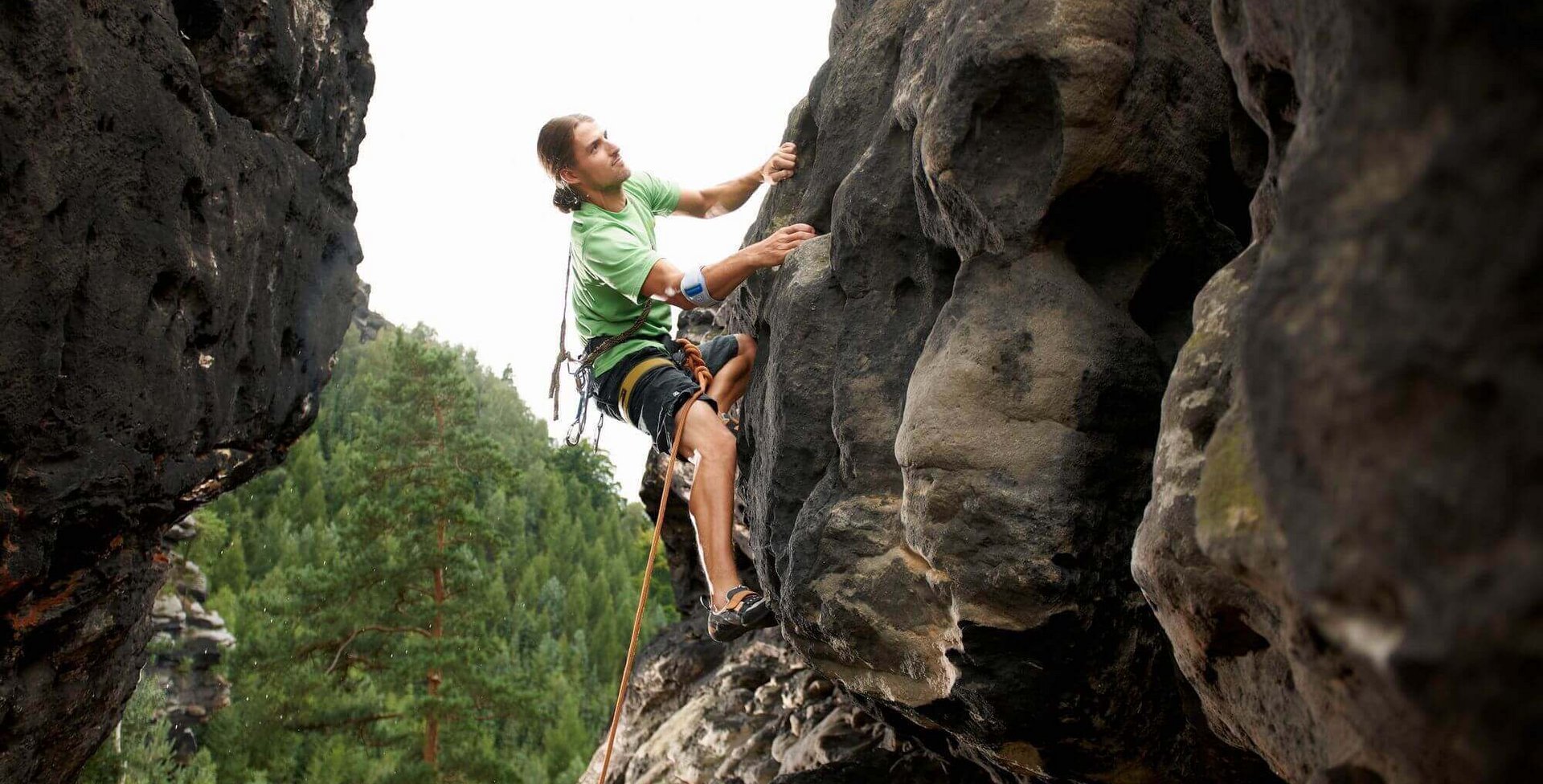 Zu sehen ist ein junger Mann, der mit Sicherung einen Felsen erklimmt. Er trägt eine EpiPoint Spange am rechten Arm, die auch Unterstützung bei einem Golfarm bietet.