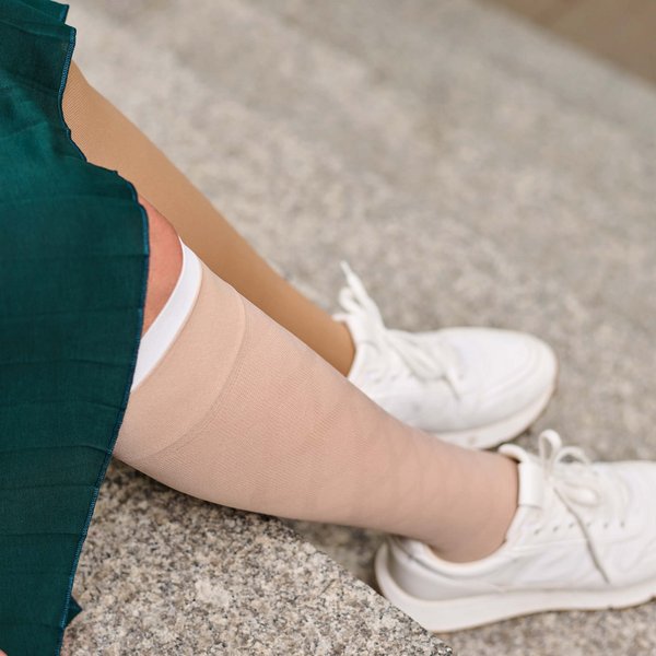 Nahaufnahme eines Beines in einem beige-farbenen Kompressionsstrumpf aus der VenoTrain Special-Linie, kombiniert mit einem grünen Kleid und weißen Sneakers. Das Bein ruht auf einer steinernen Treppenstufe.