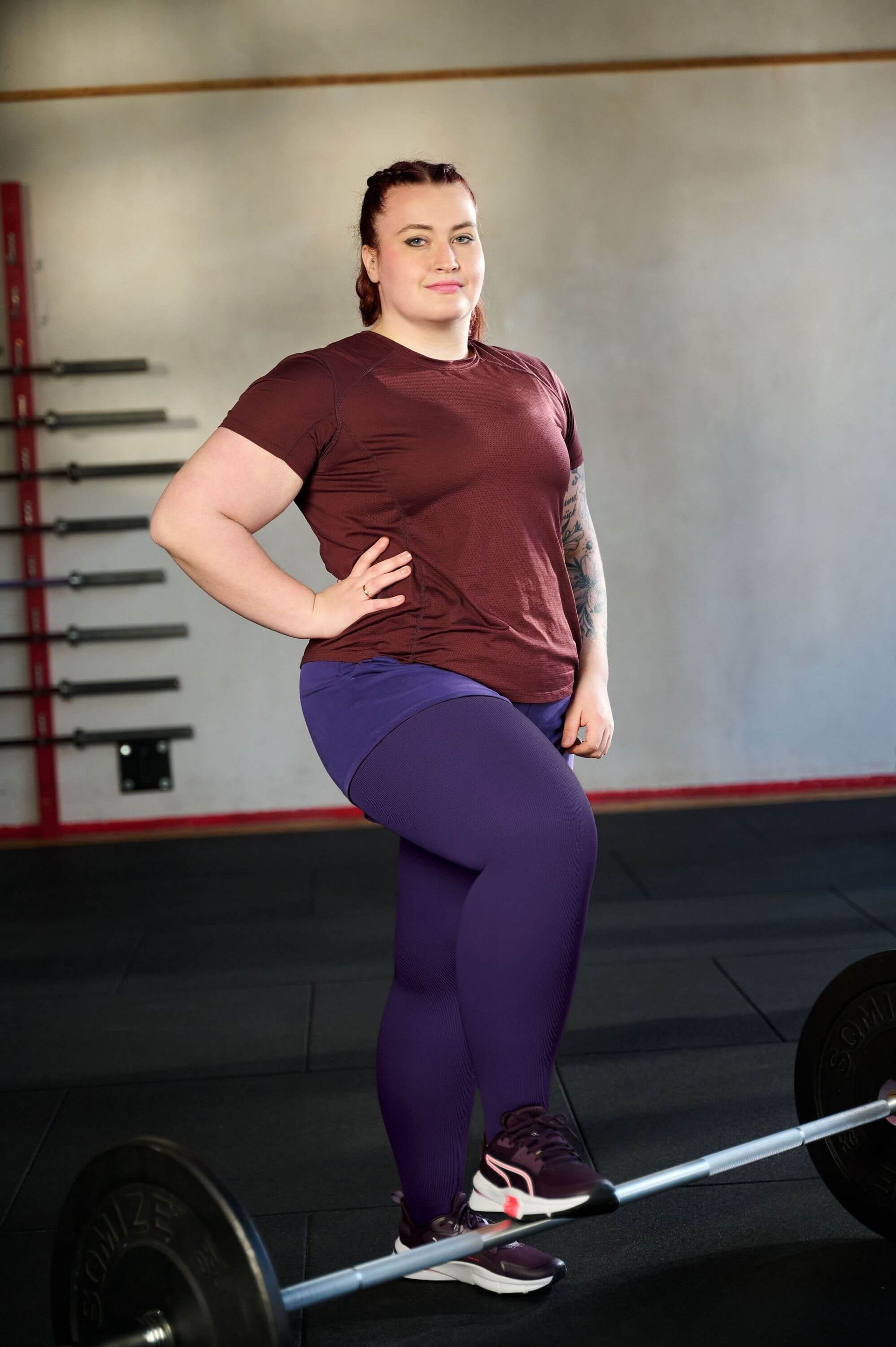 Sportlich gekleidete Frau mit rotem T-Shirt und violetter Leggings steht selbstbewusst in einem Fitnessstudio und hat ein Bein auf eine Langhantel gestützt. Im Hintergrund sind mehrere Langhantelstangen an einer Wandhalterung sichtbar.