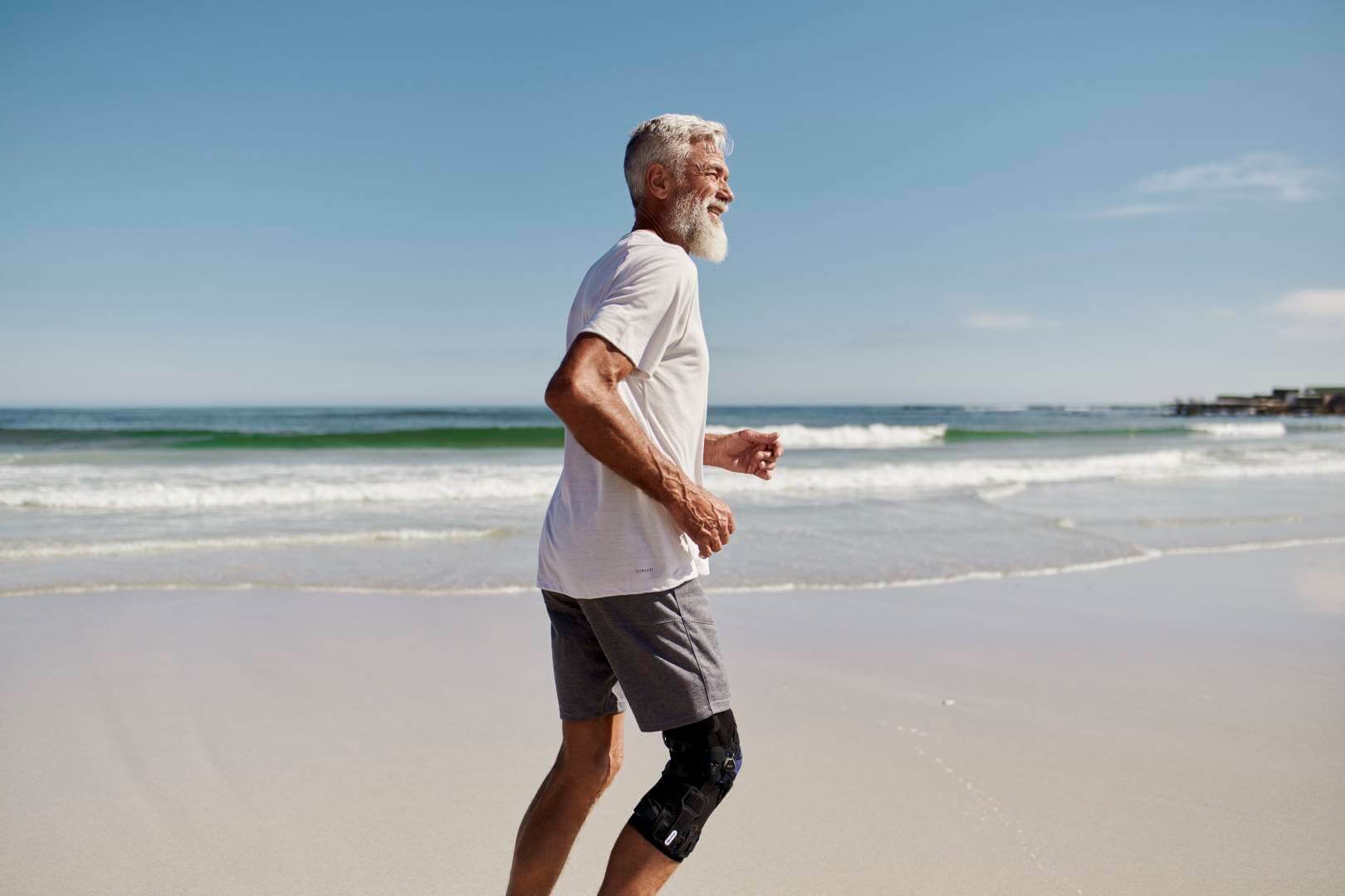 Ein älterer Herr joggt am Strand entlang. Im Hintergrund ist das Meer zu sehen. Er trägt eine GenuTrain OA von Bauerfeind am rechten Knie. Die Orthese wurde speziell für Arthrose-Patienten entwickelt.