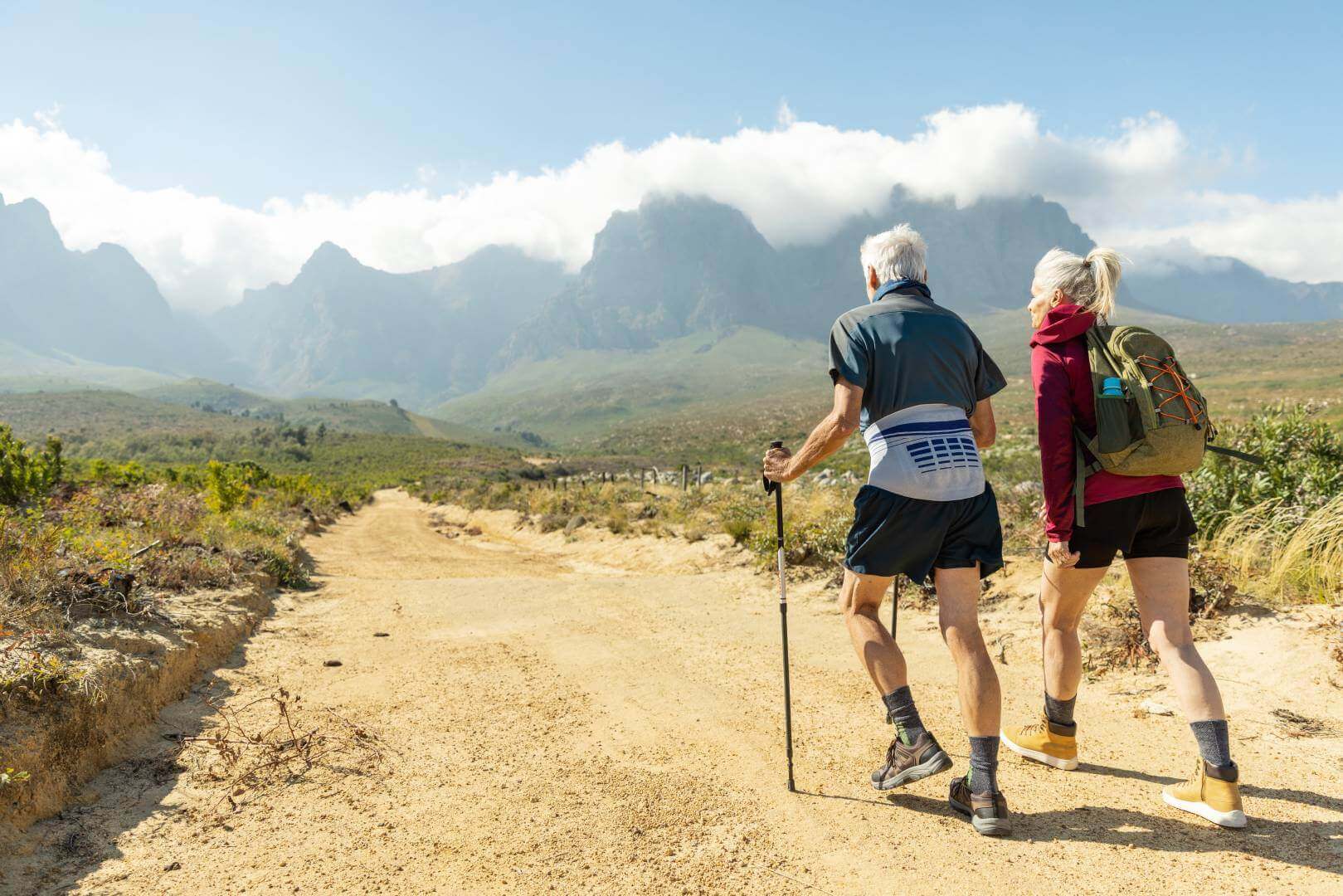 Abgebildet ist ein älteres Paar, das in einer sommerlichen Berglandschaft wandert. Zur Unterstützung nutzt der Mann Wanderstöcke und trägt eine Bauerfeind LumboLoc Rückenbandage.