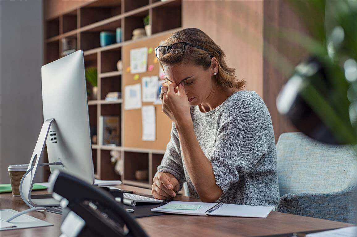 Das Bild zeigt eine gestresste Frau an ihrem Büroarbeitsplatz. Sie legt sich die Hand an die Stirn. Bewegungsmangel, z.B. bei der Arbeit, kann sich auch auf die Psyche auswirken.