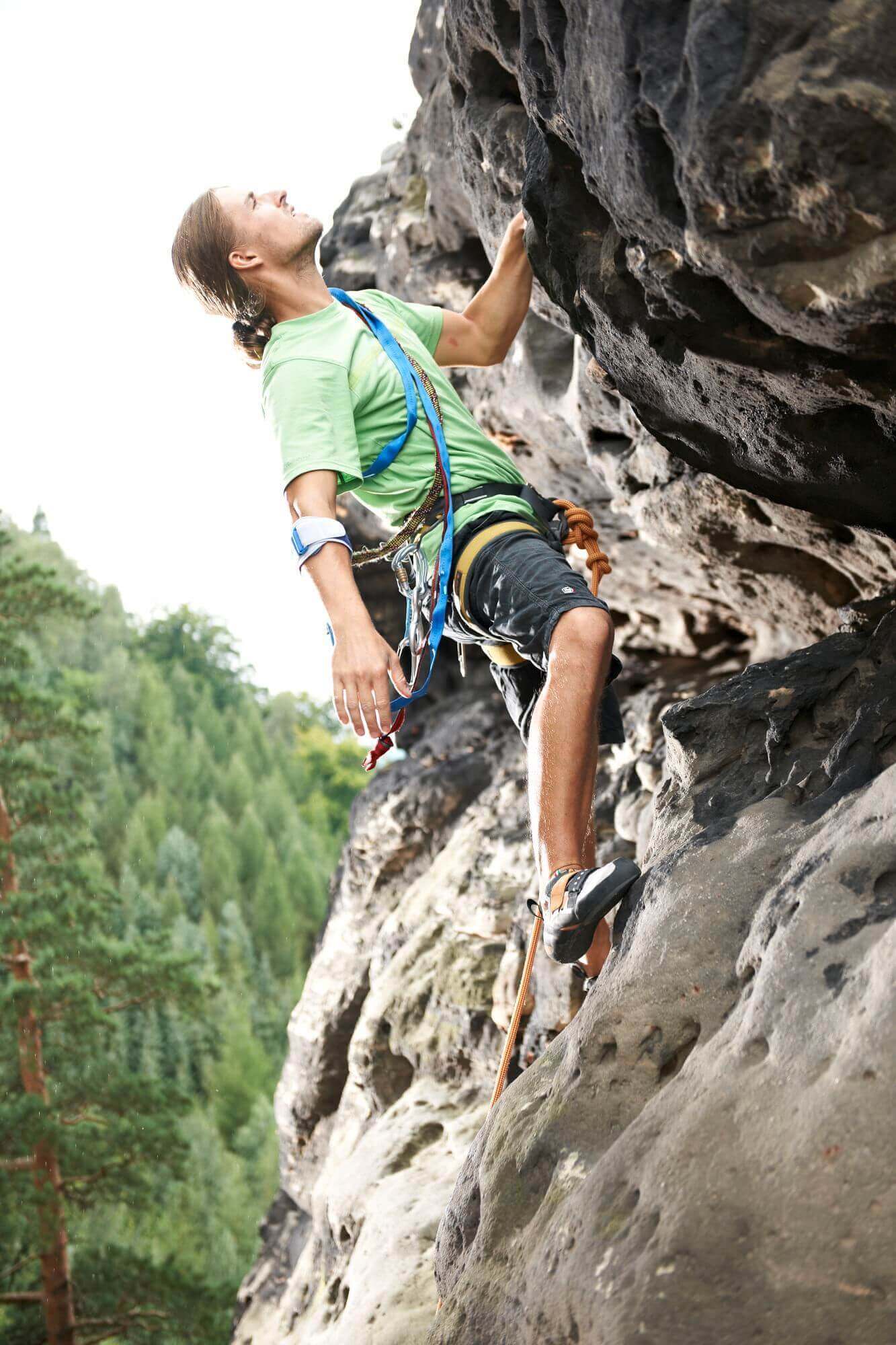 	Ein junger Mann mit Sicherung klettert einen Felsen hoch. Er trägt eine EpiPoint von Bauerfeind am rechten Arm. Die EpiPoint ist eine schmerzlindernde Epicondylitis-Spange, welche bei gereizten Sehnenansätzen angewendet wird.