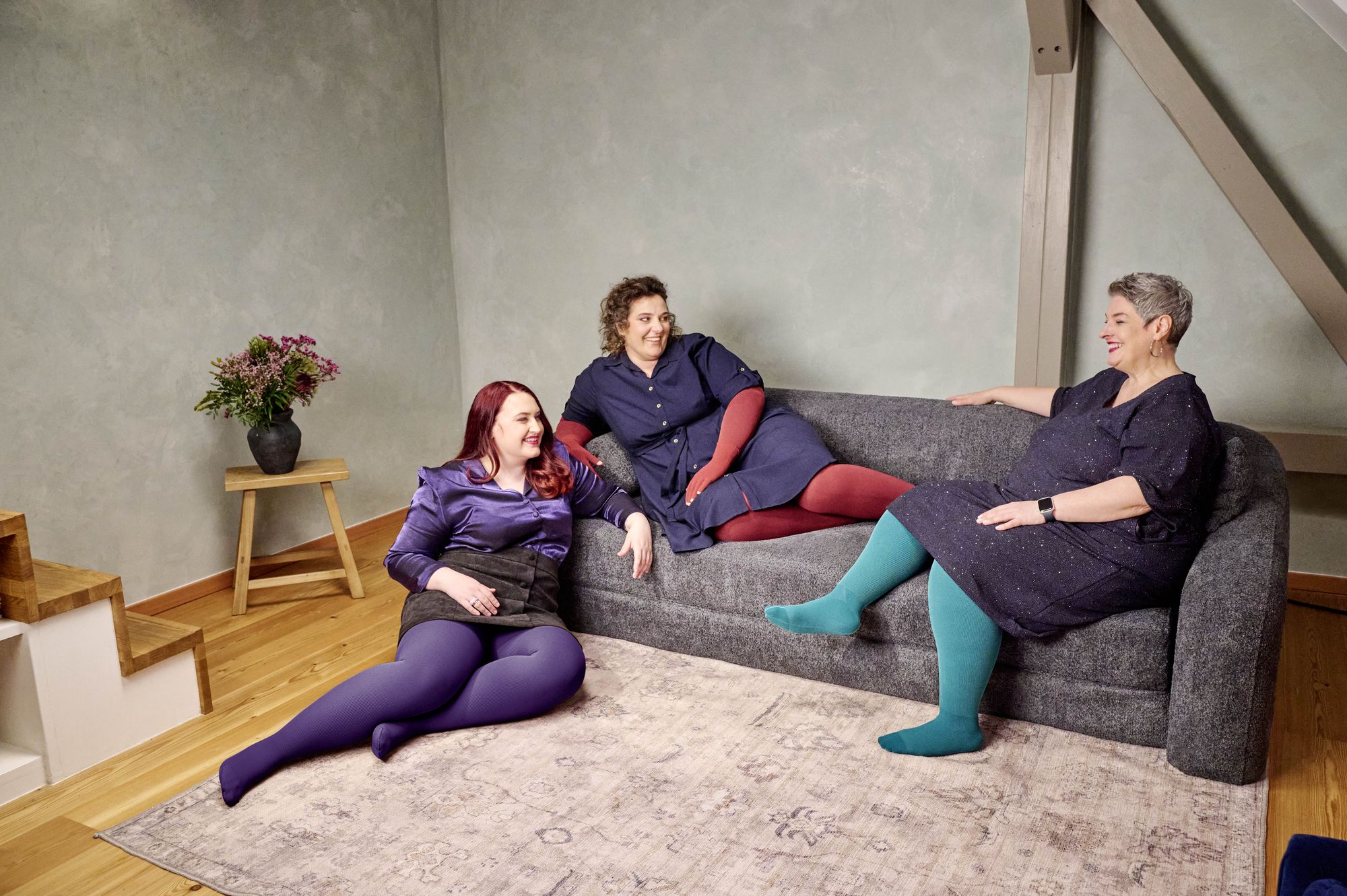 Drei Frauen sitzen und liegen entspannt auf einem dunkelgrauen Sofa in einem modern eingerichteten Wohnzimmer mit Holzfussboden. Sie tragen farbenfrohe Strumpfhosen – in Violett, Rot und Türkis – kombiniert mit dunklen Kleidern oder Röcken. Alle wirken fröhlich und unterhalten sich miteinander. Links steht ein kleiner Holztisch mit einer schwarzen Vase und einem Blumenstrauss.