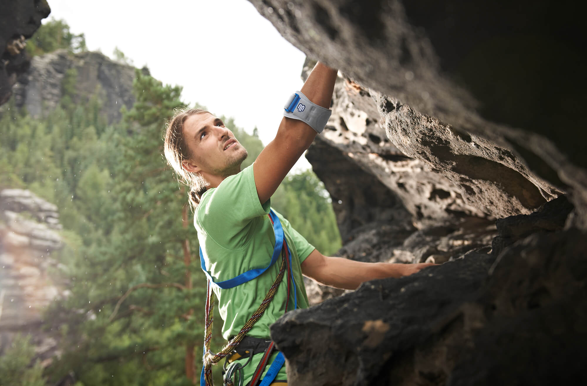 Ein junger Mann mit Kletterausrüstung klettert an einer steilen Felswand im Freien. Er trägt ein grünes T-Shirt, einen Klettergurt mit Seilen und eine Bandage am rechten Arm. Im Hintergrund sind dichte grüne Nadelbäume und Felsen zu sehen.