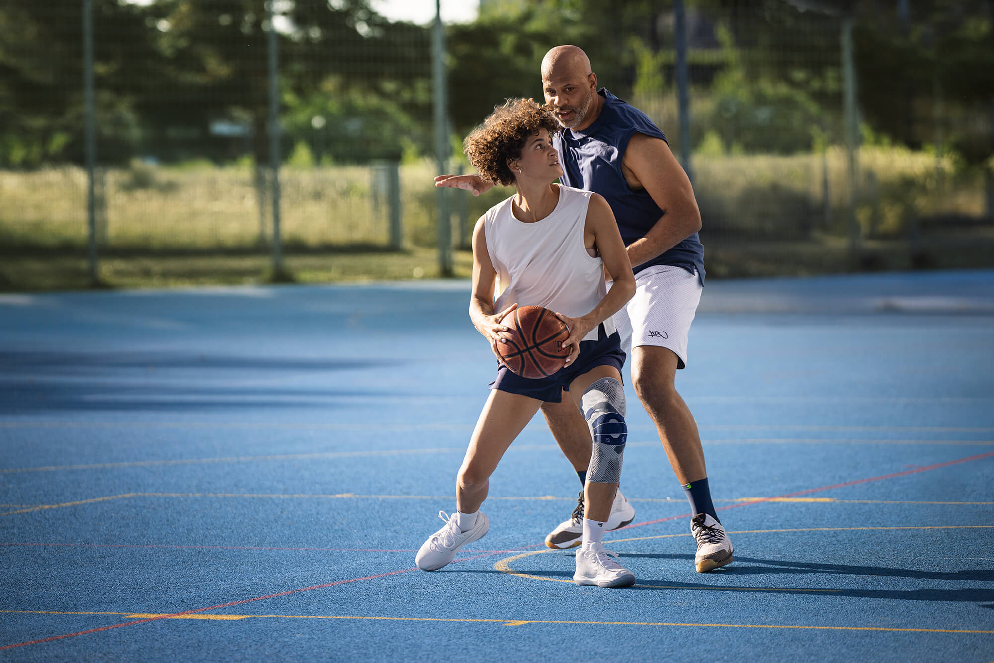 Zwei Personen spielen Basketball auf einem Aussenplatz mit blauem Boden. Die vordere Person mit weissem Trägershirt, dunklen Shorts und Kniebandage am rechten Bein hält den Ball in Spielposition, während die andere Person sie eng verteidigt.