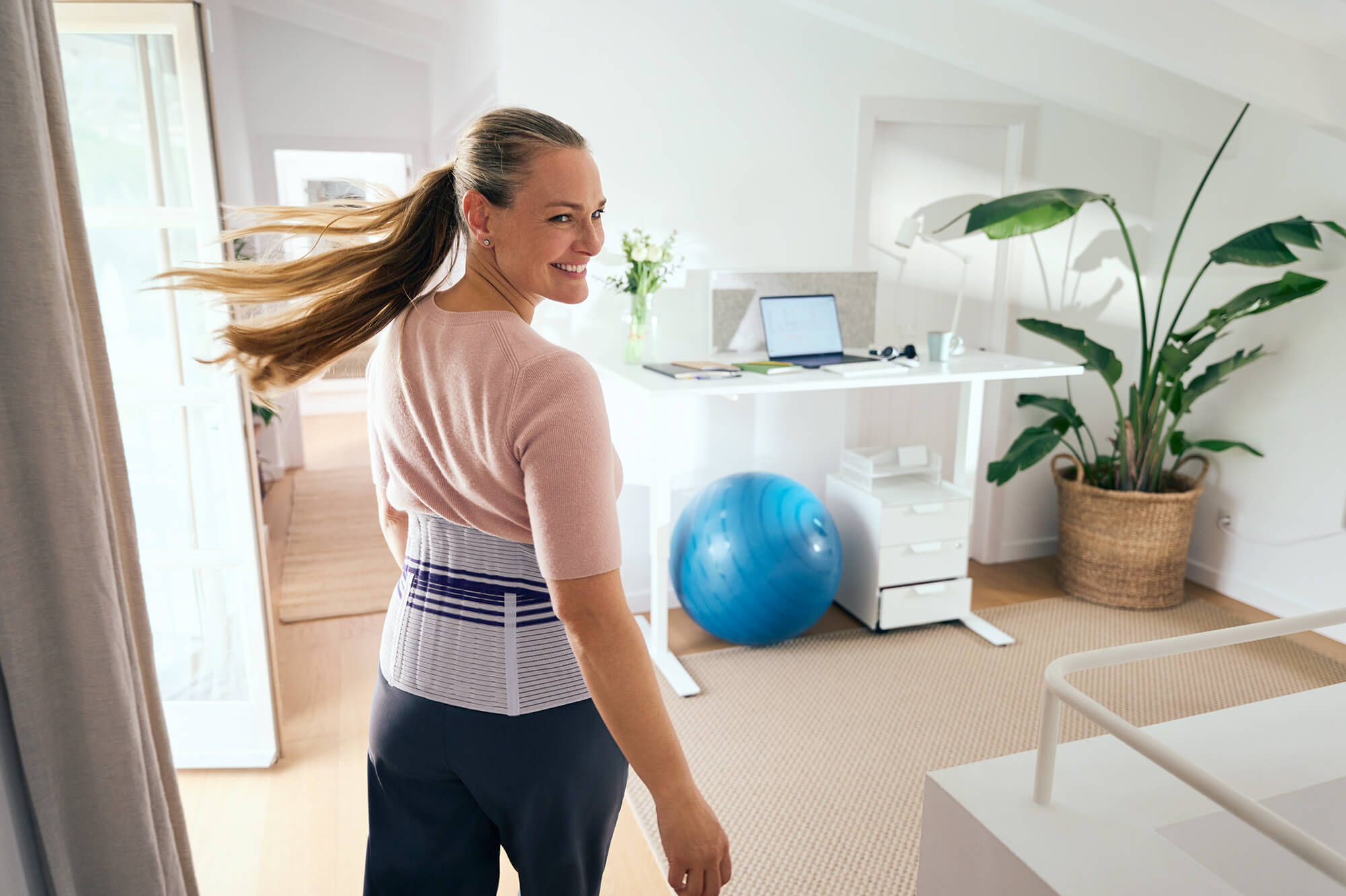 Frau mit Pferdeschwanz dreht sich lächelnd um und trägt eine graue Rückenbandage mit blauen Streifen in einem hellen Homeoffice mit Stehpult, Gymnastikball und grosser Pflanze.