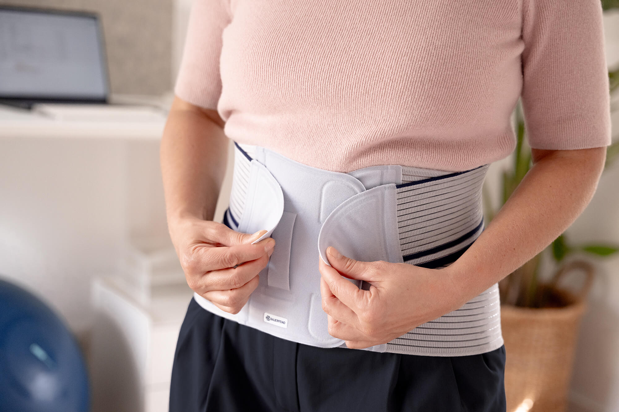 Nahaufnahme einer Frau, die mit beiden Händen eine Rückenbandage mit Klettverschluss im Bereich des unteren Rückens anlegt; im Hintergrund ein Schreibtisch mit Laptop.
