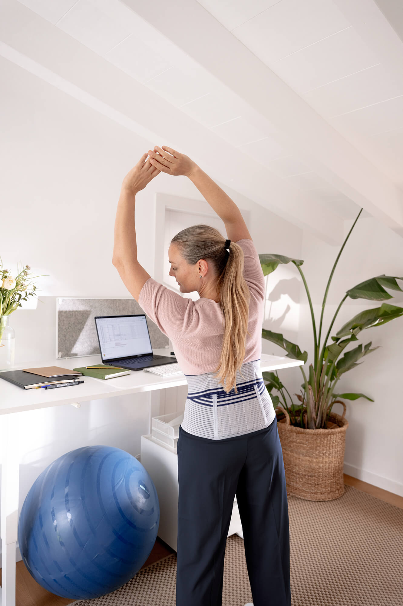 Frau mit Rückenbandage streckt sich mit erhobenen Armen vor einem Stehpult im Homeoffice; im Bild sind ein Gymnastikball, Notizbücher und eine grosse Pflanze zu sehen.