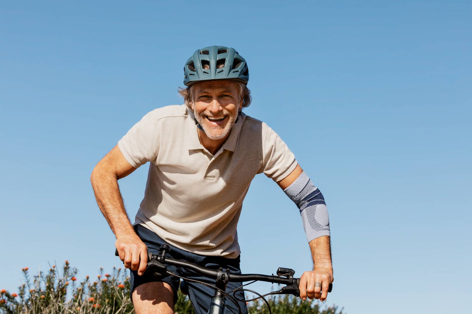 Mann mit Fahrradhelm fährt bei klarem Himmel lächelnd Fahrrad, trägt die EpiTrain Ellenbogenbandage von Bauerfeind am rechten Arm zur Unterstützung des Gelenks.